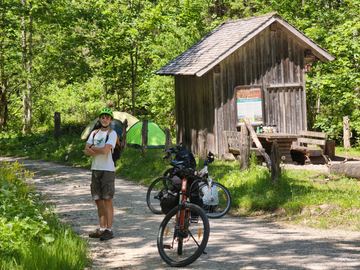 Reko in a green helmet next to loaded bikes and tents at a wooden forest shelter