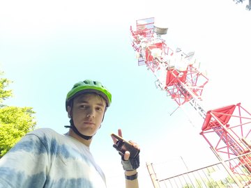 Reko in a green bike helmet holding up a walkie-talkie under a tall red and white transmitter tower