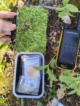 A geocache box hidden under a patch of fake grass, a walkie-talkie lying next to it on the forest floor