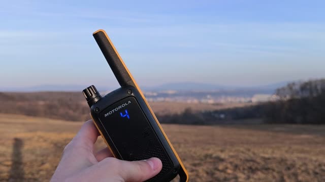 Holding up a walkie-talkie over wide meadows in the late afternoon sun, a town and hills in the distance
