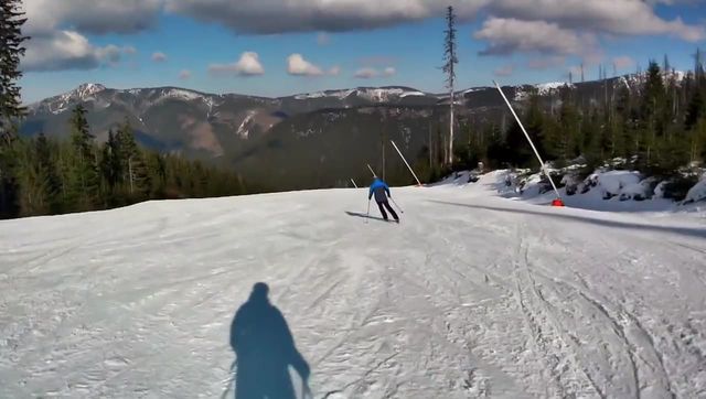 Skiing down a sunny groomed slope between spruce forests, mountains in the distance, seen from the skier's view